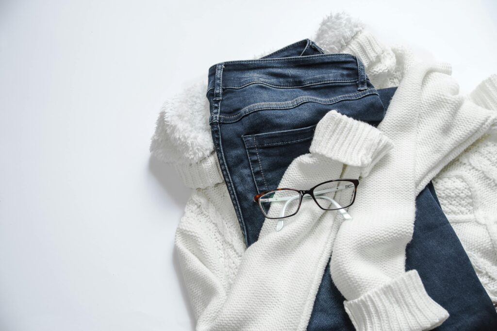 Stylish flat lay featuring denim jeans, white sweater, and eyeglasses on a bright background.